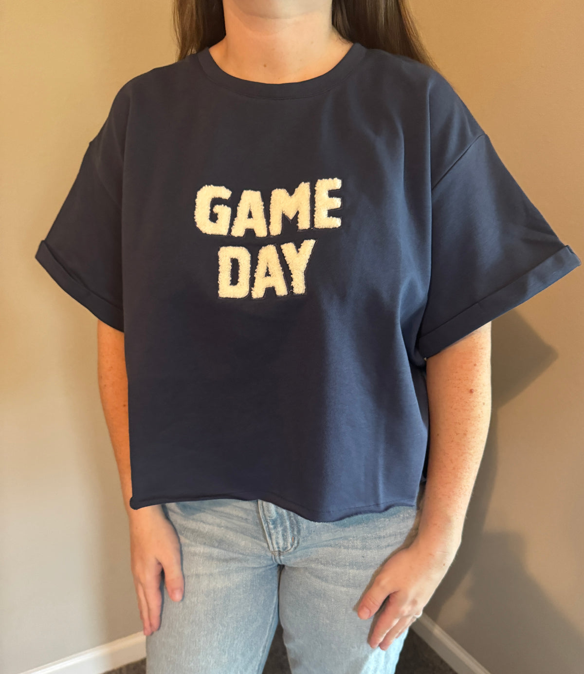 A person wearing an oversized navy blue t-shirt with 'GAME DAY' printed in cream across the chest, paired with light-wash jeans, standing against a beige wall.