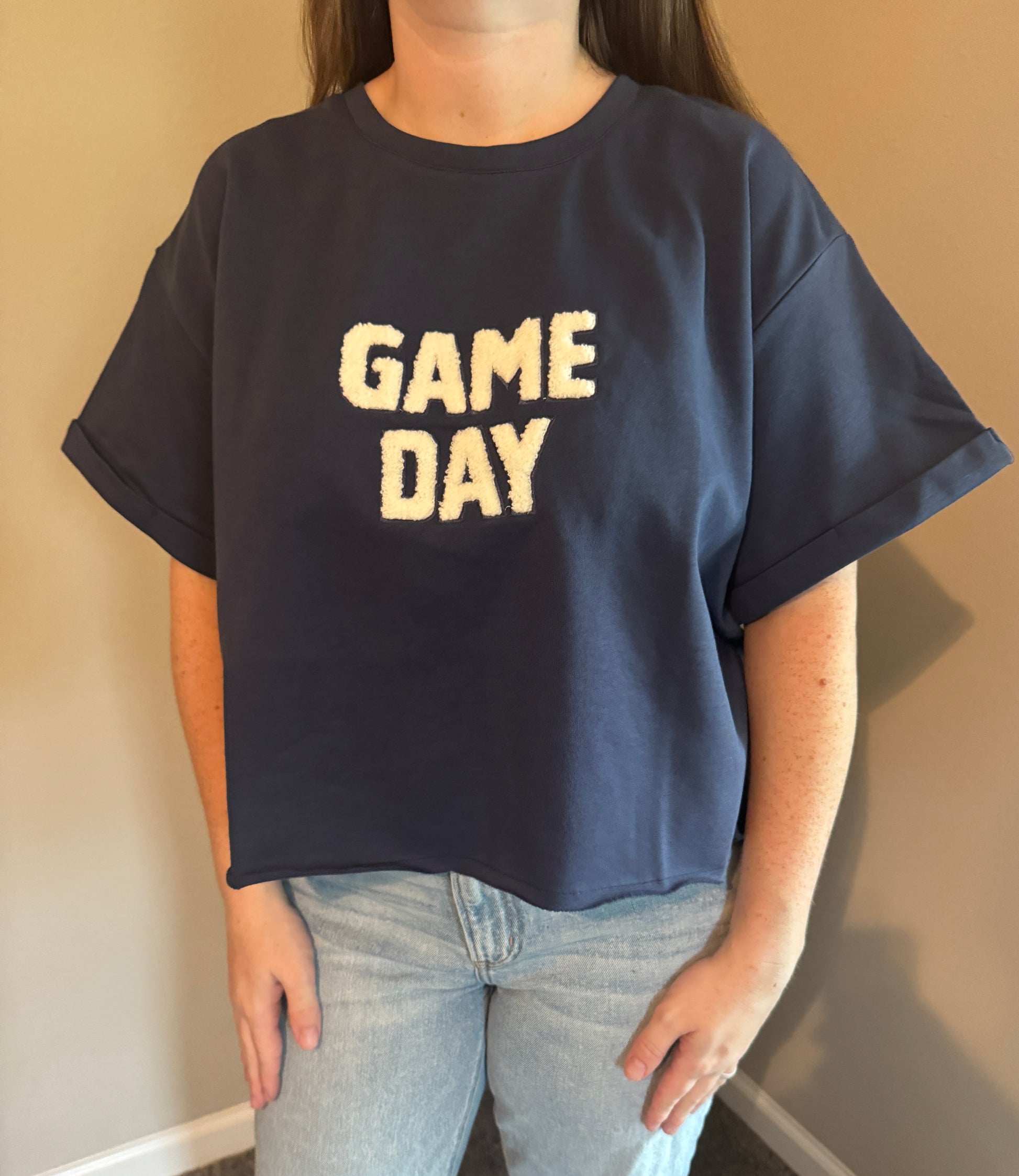 A person wearing an oversized navy blue t-shirt with 'GAME DAY' printed in cream across the chest, paired with light-wash jeans, standing against a beige wall.