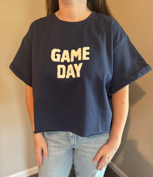 A person wearing an oversized navy blue t-shirt with 'GAME DAY' printed in cream across the chest, paired with light-wash jeans, standing against a beige wall.